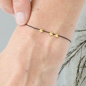 Pode incluir: Um simples bracelete de corda preta com três pingentes de estrela dourados.