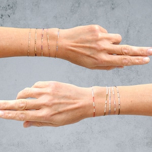 Könnte beinhalten: Ein Set aus zarten Armbändern mit kleinen Perlen in verschiedenen Farben, darunter Gold, Silber, Rot und Blau. Die Armbänder sind an einem Handgelenk übereinander getragen.