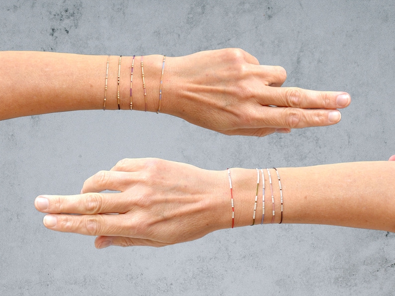 May include: A set of delicate bracelets with small beads in various colors, including gold, silver, red, and blue. The bracelets are stacked on a person's wrist.