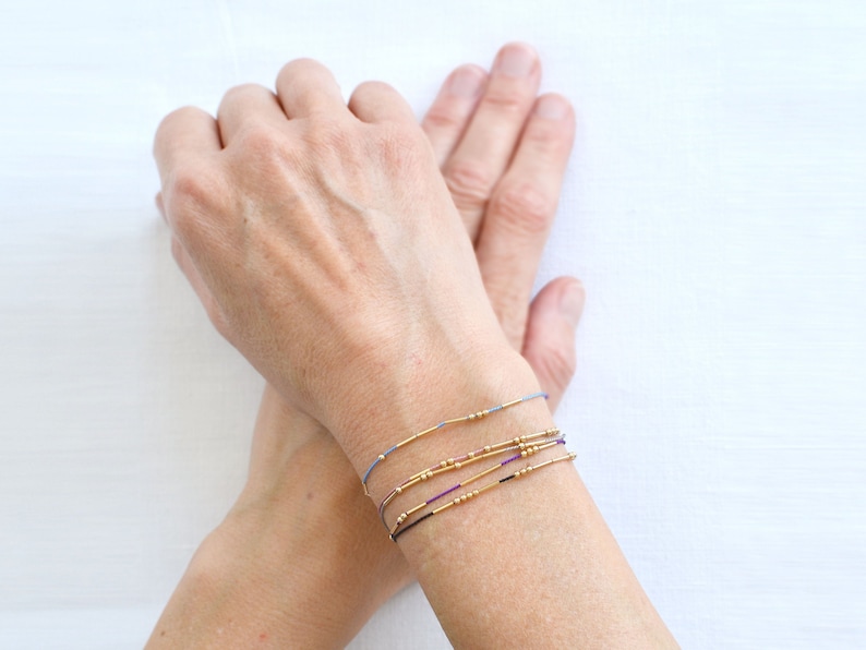 May include: A person's wrist with multiple thin bracelets made of gold beads and colored string. The bracelets are layered on top of each other.