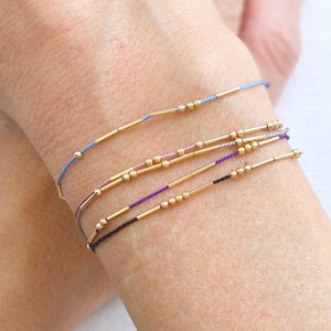May include: A set of five bracelets with gold beads and thin gold bars. The bracelets are layered on a person's wrist and are in shades of blue, purple, pink, and black.