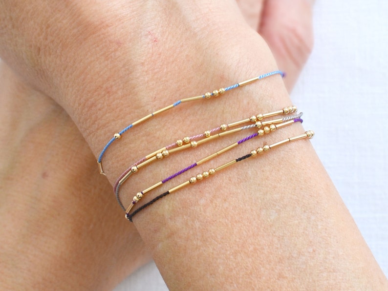 May include: A set of five bracelets with gold beads and thin gold bars. The bracelets are layered on a person's wrist and are in shades of blue, purple, pink, and black.
