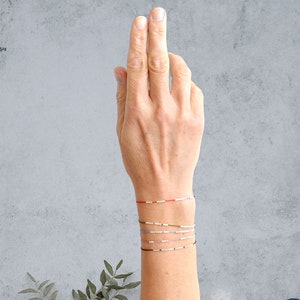 May include: A hand wearing a stack of colorful bracelets. The bracelets are made of small beads and string. The bracelets are in shades of red, white, blue, green, and gray.