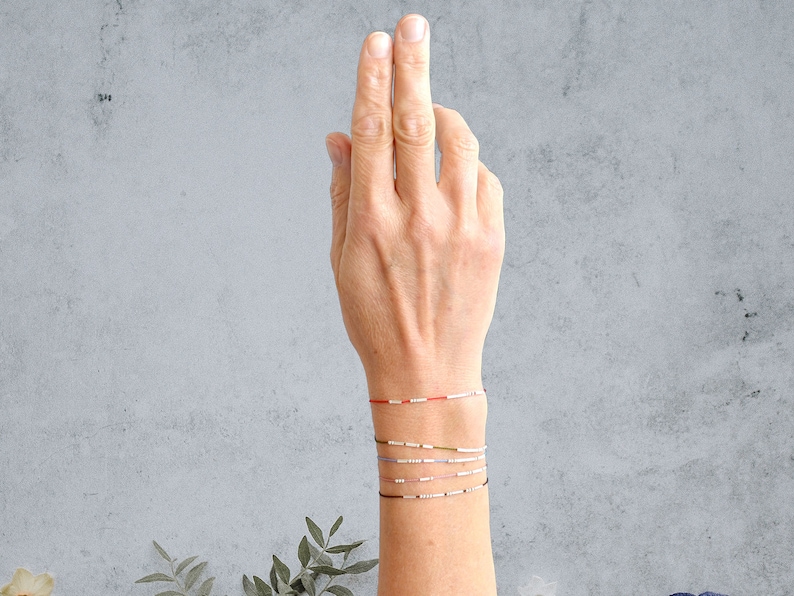 May include: A hand wearing a stack of colorful bracelets. The bracelets are made of small beads and string. The bracelets are in shades of red, white, blue, green, and gray.