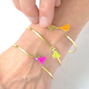 May include: Gold-toned metal bracelets with colorful tassel accents. The bracelets feature a simple, open design and are adorned with small, round charms. Tassels are in bright pink, yellow, and orange. The bracelets are worn on a wrist.
