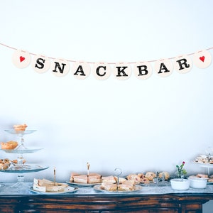 May include: A white wall with a red and white string banner that reads "SNACKBAR" with red hearts on each end. A table with a white tablecloth is set up with a variety of snacks and desserts, including pastries, sandwiches, and cookies.