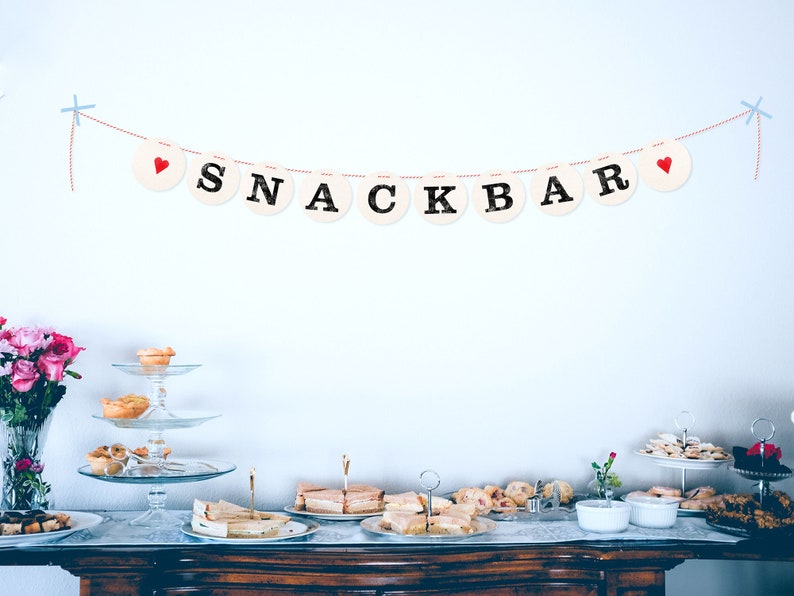 May include: A white wall with a red and white string banner that reads "SNACKBAR" with red hearts on each end. A table with a white tablecloth is set up with a variety of snacks and desserts, including pastries, sandwiches, and cookies.