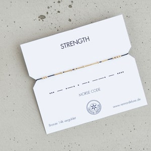 May include: A white card with the word "STRENGTH" printed in black. The card also features a blue string bracelet with gold beads, and the Morse code for "STRENGTH" printed below the bracelet. The card also includes the text "MORSE CODE" and the website "www.renna-deluxe.de".