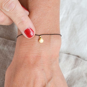 Puede incluir: Un delicado brazalete de cuerda negra con un pequeño dije de concha dorada.
