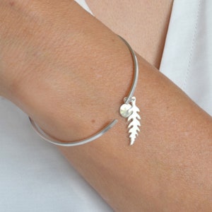 May include: A silver bangle bracelet with a delicate fern charm hanging from it. The charm is made of silver and has a small, round bead at the top.