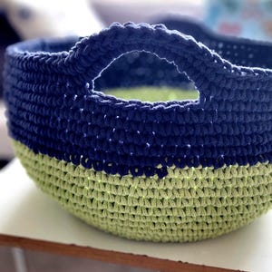 May include: A crocheted storage basket with a blue and green color scheme. The basket has a single handle and is made of thick cotton rope.