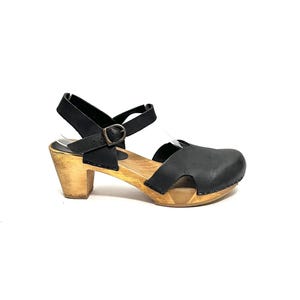 May include: Black leather clogs with a wooden sole and a 2-inch heel. The clogs feature an adjustable ankle strap with a metal buckle. The shoe has a closed toe design and a natural wood finish on the heel and sole.