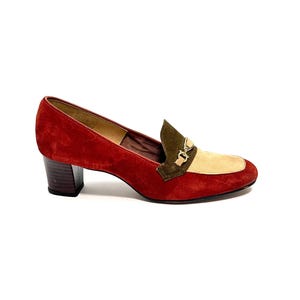 May include: A red suede heeled loafer with a square toe. The shoe features a tan and olive green accent on the vamp, with a gold-tone buckle. The heel is a dark brown block heel.