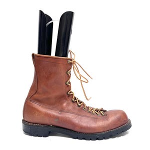 May include: A brown leather lace-up boot with a black rubber lug sole and metal eyelets. The boot features a high-cut ankle design, tan laces, and a black plastic boot shaper inserted into the opening to maintain the shape of the leather upper.