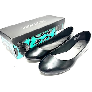 May include: A pair of black, glossy ballet flats next to a box. The shoes have a rounded toe and a low heel. The box is black with the words "LOWER EAST SIDE" on top. The box has a teal and black floral design.