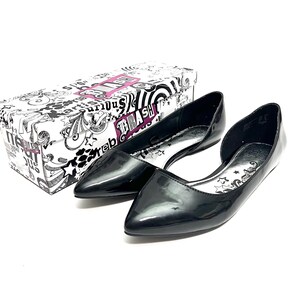 May include: Pair of black patent leather flats with pointed toes. The shoes have a low-cut vamp and a silver insole with star and text details. A matching box with a black and white graphic design is in the background.