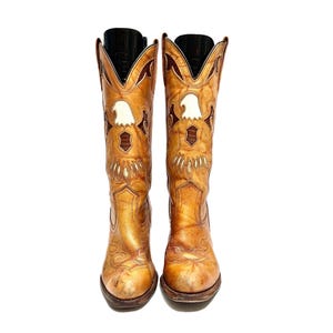 Peut inclure: Une paire de bottes de cowboy en cuir bronzé avec un motif d'aigle. Les bottes ont un bout pointu et une tige haute. L'aigle est blanc avec des accents marron et bronzé. Les bottes sont de style western classique.