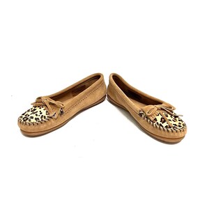 Puede incluir: Un par de mocasines de ante marr&oacute;n con un dise&ntilde;o de estampado de leopardo en la punta. Los mocasines tienen un detalle de borla y un aspecto suave y c&oacute;modo.