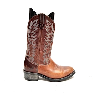 Puede incluir: Una bota de vaquero de cuero marrón con costuras decorativas y suela negra. La bota tiene un aspecto desgastado y un tacón apilado. La caña presenta un patrón decorativo cosido. La bota está diseñada para montar a caballo y actividades al aire libre.