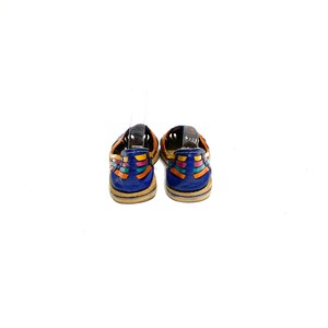 May include: A pair of colourful leather sandals. The sandals have a blue heel and are decorated with woven orange, yellow, and turquoise straps. The soles are a light tan colour. The sandals are displayed against a white background.