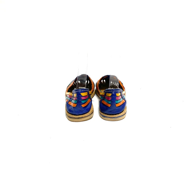 May include: A pair of colourful leather sandals. The sandals have a blue heel and are decorated with woven orange, yellow, and turquoise straps. The soles are a light tan colour. The sandals are displayed against a white background.
