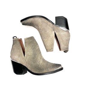 May include: A pair of grey ankle boots with a textured pattern. The boots have a V-cut design on the sides, black block heels, and silver-toned metal accents on the toe and heel. The boots are approximately 15 cm in height.