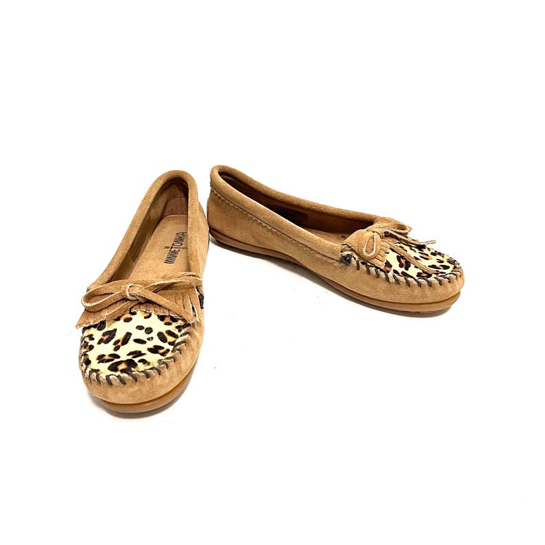 Puede incluir: Un par de mocasines de ante marr&oacute;n con un dise&ntilde;o de estampado de leopardo en la punta. Los mocasines tienen un detalle de flecos y un lazo en la parte superior.