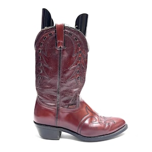 Könnte beinhalten: Ein dunkelburgunderfarbener Leder-Cowboystiefel mit strukturiertem Schaft, Ziernähten und Ausschnitten, einem glatten Vorderblatt mit flammenartiger Stickerei, einem schwarzen Blockabsatz und einer schwarzen Sohle. Enthält einen schwarzen Kunststoff-Schuhspanner.