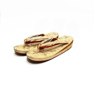 May include: A pair of traditional Japanese sandals, known as geta, with a cream-colored fabric upper featuring a gold and white floral pattern. The sandals have a red leather strap and a wooden base.