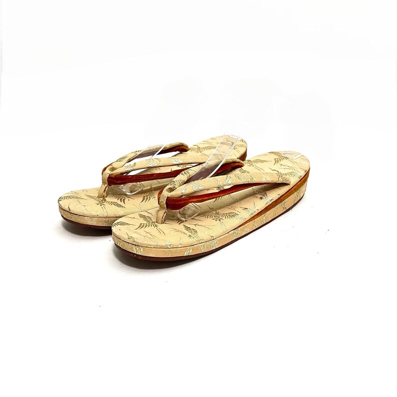 May include: A pair of traditional Japanese sandals, known as geta, with a cream-colored fabric upper featuring a gold and white floral pattern. The sandals have a red leather strap and a wooden base.