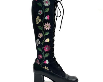 May include: Black knee-high boot with floral embroidery. The boot has a lace-up design and a block heel. The embroidered flowers are in pink, green, and white. The heel measures approximately 6.35 cm.