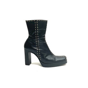 Pode incluir: Botas de tornozelo de couro preto com salto grosso e bico quadrado. As botas têm um detalhe de costura decorativa ao longo do eixo.