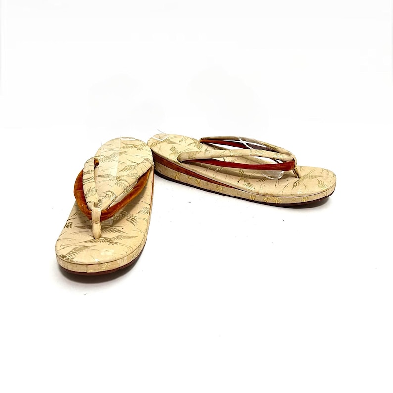 May include: A pair of traditional Japanese sandals, known as geta, with a cream-colored base and red straps. The sandals are decorated with a delicate pattern of gold leaves.