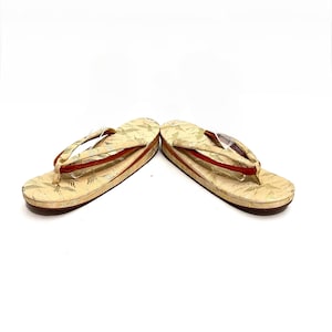 May include: A pair of traditional Japanese sandals, known as geta, with a cream-colored base and gold and red accents. The sandals have a simple design with a single strap across the foot.
