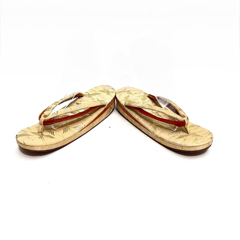 May include: A pair of traditional Japanese sandals, known as geta, with a cream-colored base and gold and red accents. The sandals have a simple design with a single strap across the foot.