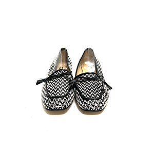 Op de afbeelding: Een paar zwart-witte geweven loafers met een zigzagpatroon. De schoenen hebben zwarte leren randen en kleine strikjes op de wreef. De schoenen staan tegen een witte achtergrond.