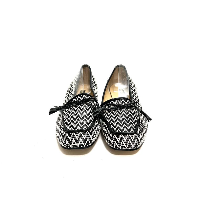 Op de afbeelding: Een paar zwart-witte geweven loafers met een zigzagpatroon. De schoenen hebben zwarte leren randen en kleine strikjes op de wreef. De schoenen staan tegen een witte achtergrond.