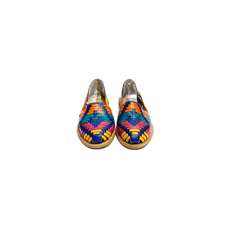May include: A pair of colourful woven leather huarache sandals. The sandals feature a vibrant pattern of blue, orange, pink, yellow, and teal. The soles are a light brown colour. The shoes are displayed on a white background.