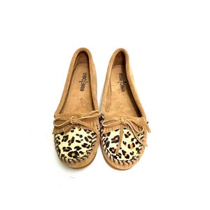 Puede incluir: Un par de mocasines de ante marr&oacute;n con detalles de estampado de leopardo en la puntera. Los mocasines tienen un detalle de flecos y el nombre de la marca "Minnetonka" est&aacute; impreso en la plantilla.