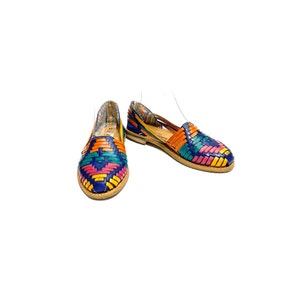 May include: A pair of colourful woven leather flats. The shoes feature a vibrant pattern of blue, orange, pink, yellow, and green. The soles are a light brown colour, and the shoes are set against a white background.