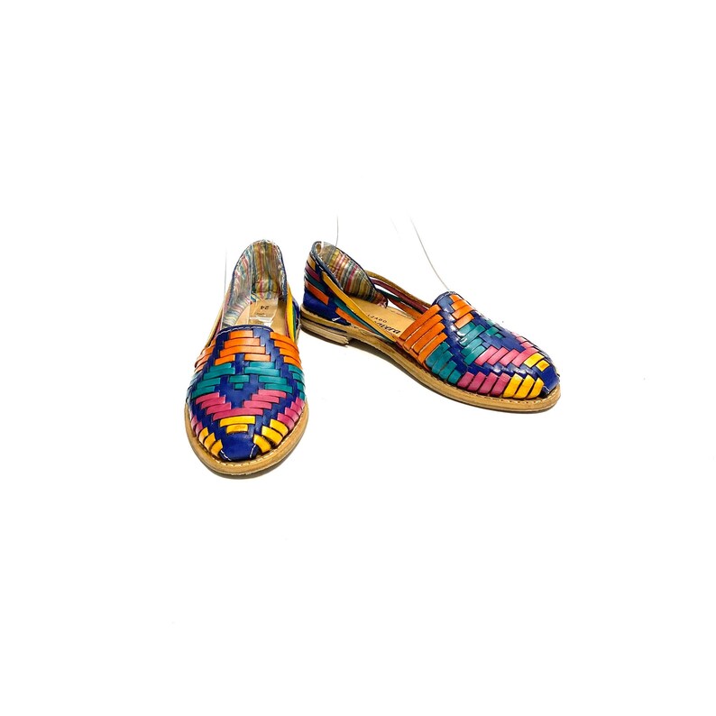 May include: A pair of colourful woven leather flats. The shoes feature a vibrant pattern of blue, orange, pink, yellow, and green. The soles are a light brown colour, and the shoes are set against a white background.
