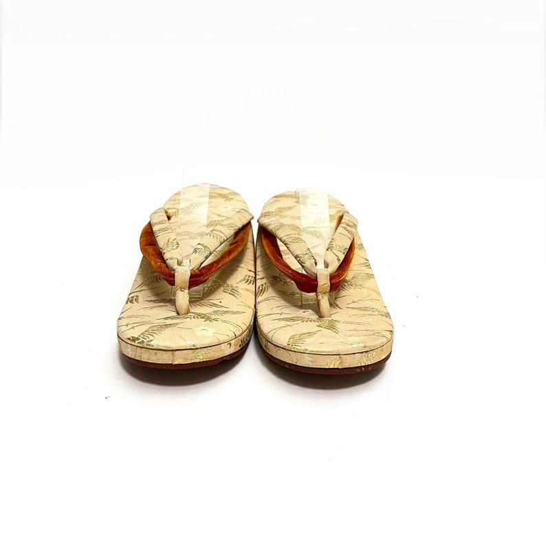 May include: A pair of white Japanese sandals with gold and orange accents. The sandals have a traditional design with a raised platform and a thong strap.