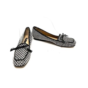 Op de afbeelding: Een paar zwart-witte geweven loafers met een vierkante neus en een kleine zwarte kwast. De schoenen hebben een zwarte bies en een lage hak. De schoenen staan op een witte achtergrond.