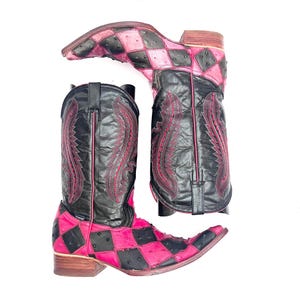 May include: A pair of cowboy boots featuring a black and hot pink checkered pattern on the foot and a solid black shaft. The shaft is decorated with intricate dark pink stitching in a flame-like design. The boots have a pointed toe and a heel.
