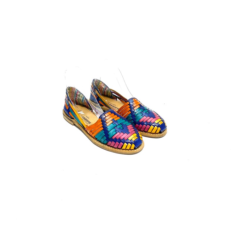 May include: A pair of colourful woven leather huarache flats. The shoes feature a vibrant pattern of blue, orange, pink, and yellow leather strips. The soles are a light brown colour, and the shoes are set against a white background.