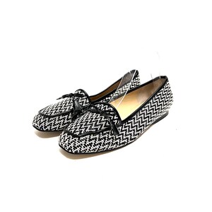 Op de afbeelding: Een paar zwart-witte geweven loafers met een zigzagpatroon. De schoenen hebben zwarte leren randen en kleine strikjes op de wreef. De schoenen staan tegen een witte achtergrond.