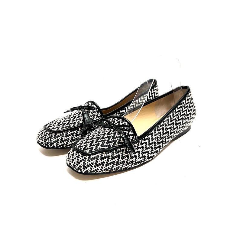 Op de afbeelding: Een paar zwart-witte geweven loafers met een zigzagpatroon. De schoenen hebben zwarte leren randen en kleine strikjes op de wreef. De schoenen staan tegen een witte achtergrond.