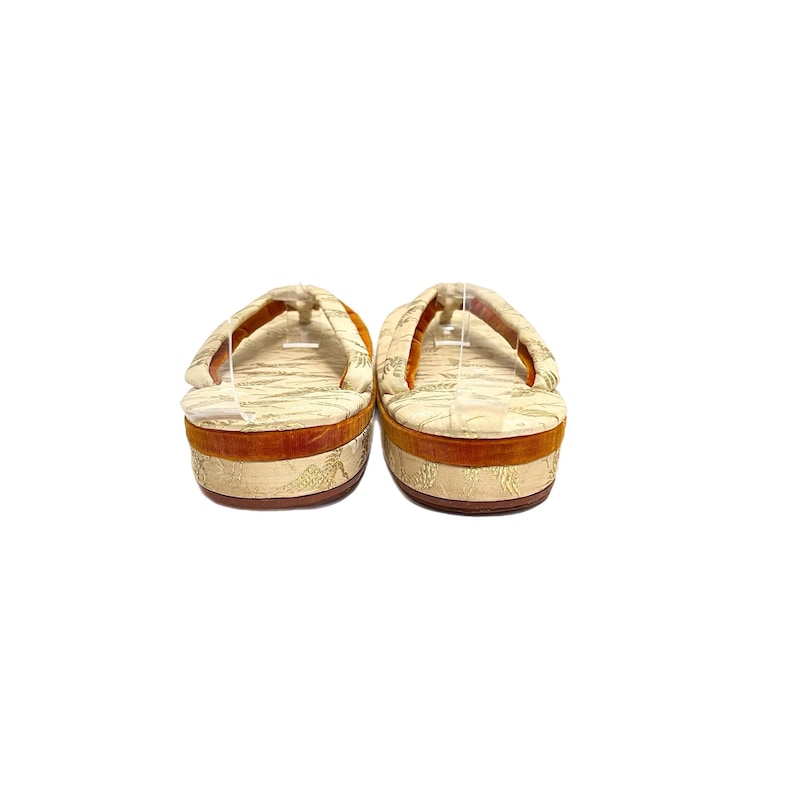 May include: A pair of traditional Japanese sandals, known as geta, with a white and gold patterned fabric upper and a brown wooden base.