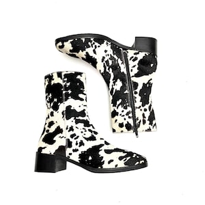 May include: A pair of ankle boots with a cowhide pattern in black and white. The boots have a block heel and a side zipper. The boots are set against a white background.
