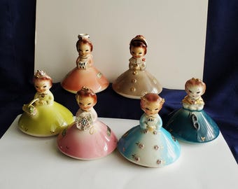 May include: A collection of six vintage ceramic figurines of young girls in dresses. The figurines are in various colors, including pink, yellow, blue, and teal. Each figurine has a unique hairstyle and holds different objects, such as a basket or flowers.